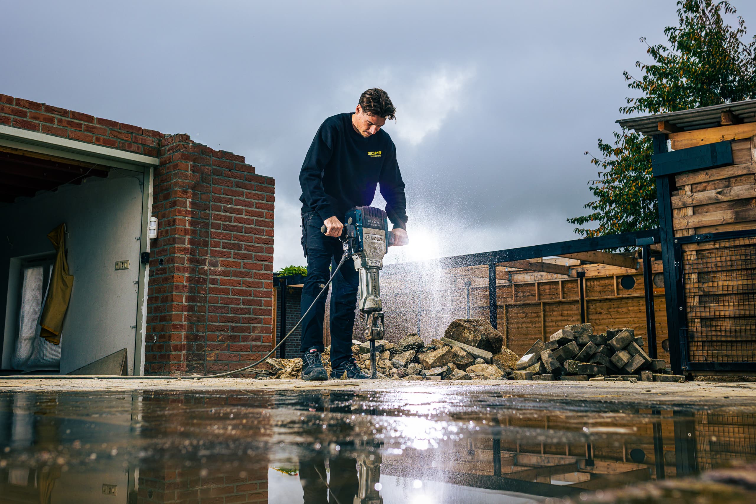 Vakman breekt met een sloophamer de bestrating op naast een garage, het natte plaveisel weerspiegelt het licht